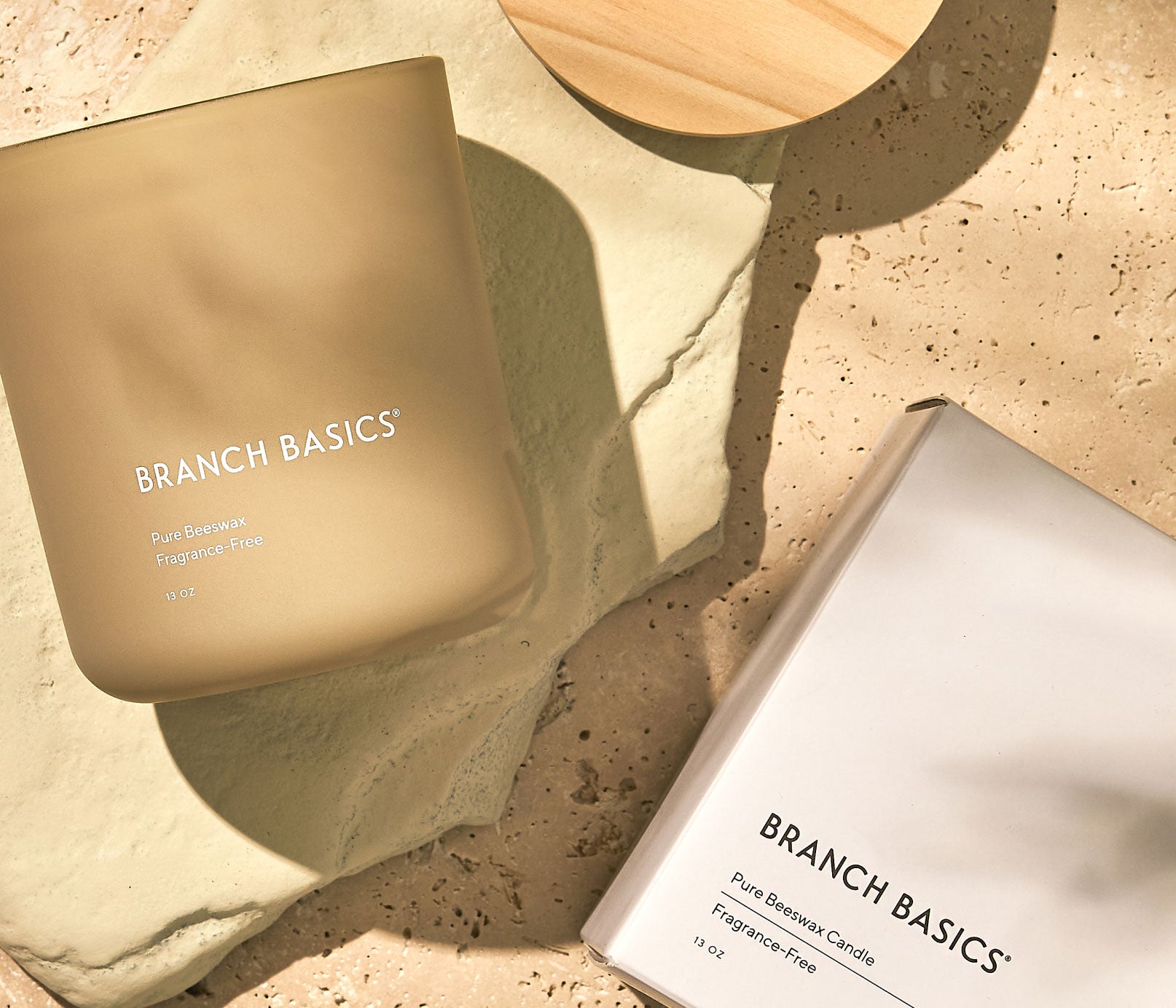 A Branch Basics 100% Beeswax Candle,  laying on its side with light refracting through the glass jar, beside a wooden lid and original packaging, displayed on a neutral stone background. - View 3
