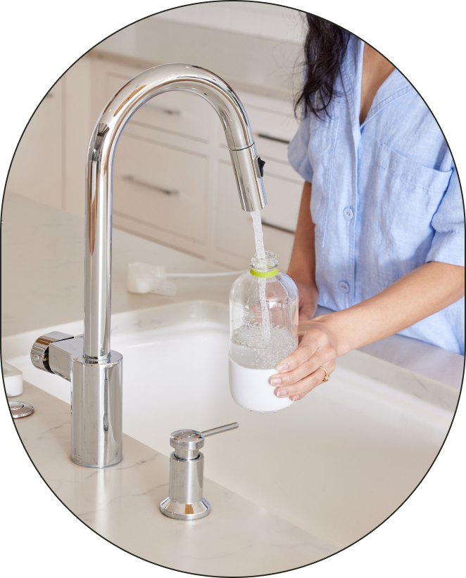 A person standing at a running sink, adding water to the water fill-line of their reusable Glass All-Purpose bottle.

